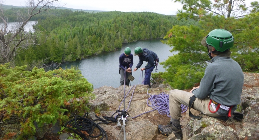 In the foreground, one person wearing safety gear rests on a rock, watching two others on the edge of a cliff. They are also wearing safety gear and are secured by ropes. One appears to be an instructor, giving direction to a student.